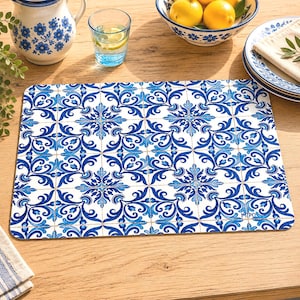 May include: A rectangular placemat with a blue and white tile pattern. The design features floral and scroll motifs in varying shades of blue against a white background. The placemat is on a wooden table, with a pitcher, glass, and bowl visible.