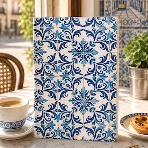 Può includere: Un taccuino con un motivo a piastrelle blu e bianche, che ricorda le tradizionali piastrelle portoghesi azulejo, è appoggiato su un tavolo. Una tazza di caffè, un pasticcino e penne con disegni coordinati sono anch'essi presenti. La scena è all'aperto.