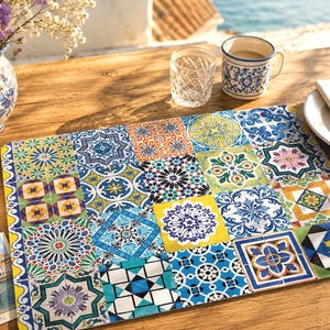 Peut inclure: Un set de table décoratif avec un motif patchwork de carreaux colorés bleus, jaunes, oranges et verts. Il est posé sur une table en bois, avec une tasse, un verre et une assiette à proximité. Le design évoque le style méditerranéen.