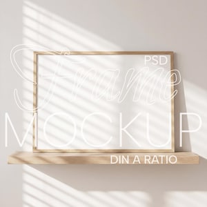 以下が含まれることがあります： 白い手書きフォントで「Frame Mockup」と「PSD DIN A RATIO」の文字が入った長方形の木製フレームモックアップ。フレームは白い壁を背景に、明るい色の木製の棚の上に置かれ、ブラインドの影が落ちています。