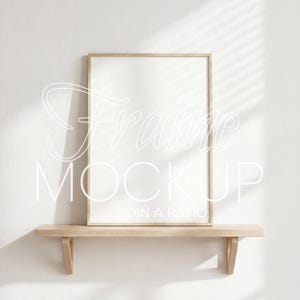 May include: A rectangular frame mockup with a light wood finish, resting on a matching wooden shelf. The frame has a white interior, and the text "Frame Mockup" is overlaid in a script font. The background is a white wall with shadows.