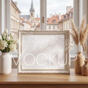 May include: A rectangular picture frame with an ornate, textured gold and white border. The frame is positioned on a wooden surface between a white vase filled with white flowers and a beige vase containing tall, dried pampas grass stalks.