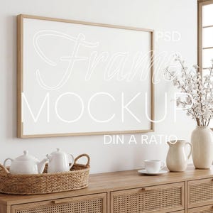 Puede incluir: Un marco de fotos rectangular horizontal con acabado de madera clara montado en una pared. El marco contiene un inserto blanco con el texto Frame MOCKUP PSD DIN A RATIO. Debajo del marco hay un aparador de madera con una bandeja tejida que contiene dos teteras blancas, una jarra de cerámica blanca y una taza de té blanca con platillo, junto a un jarrón lleno de flores blancas.