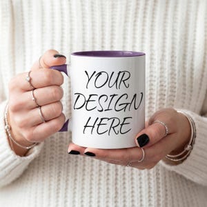 K&ouml;nnte beinhalten: Wei&szlig;er Keramikbecher mit lila Innenraum und Henkel, mit dem schwarzen Text "YOUR DESIGN HERE". Der Becher wird von einer Person gehalten, die Ringe und Armb&auml;nder tr&auml;gt. Der Becher hat eine Standardgr&ouml;&szlig;e.