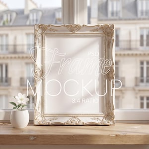 May include: A white ornate picture frame with gold accents stands on a wooden surface. The frame has a blank interior and the text "Frame Mockup 3:4 Ratio" is visible. A small white vase with a white flower is to the left.