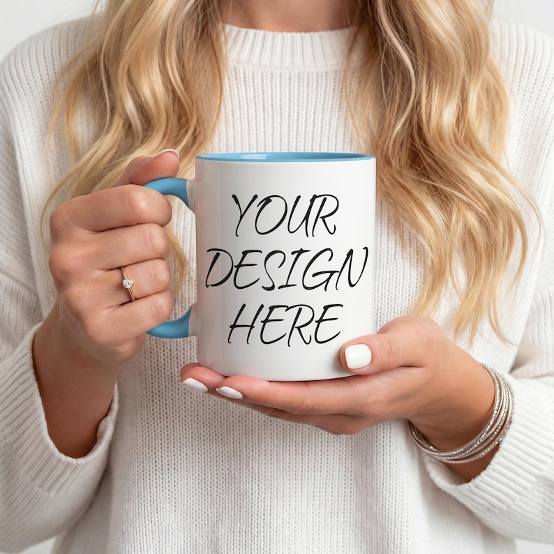 K&ouml;nnte beinhalten: Wei&szlig;er Keramikbecher mit hellblauem Innenraum und Henkel. Der Becher tr&auml;gt den Text "YOUR DESIGN HERE" in Schwarz. Der Becher wird von einer Person gehalten, die einen wei&szlig;en Pullover tr&auml;gt.