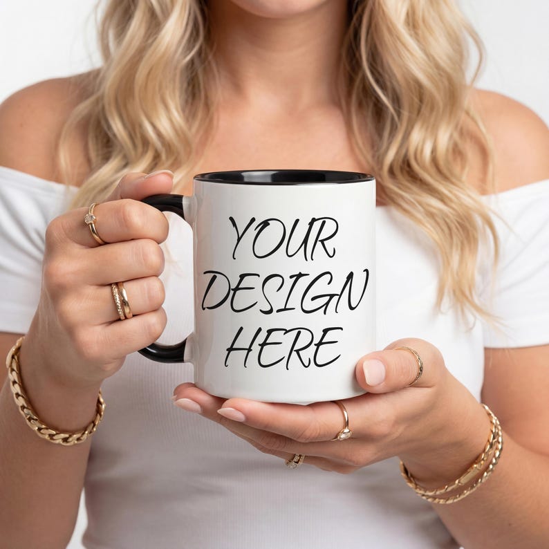 K&ouml;nnte beinhalten: Wei&szlig;er Keramikbecher mit schwarzem Innenraum und Henkel. Der Becher tr&auml;gt den Text "YOUR DESIGN HERE" in schwarzer, handschriftlicher Schrift. Der Becher wird von einer Person gehalten, die Goldschmuck tr&auml;gt.