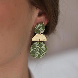 May include: Geometric earrings in green and gold. The earrings comprise a green circle, a gold half-circle, and a green circle. The green circles have a white floral pattern. The earrings are suspended from a person's ear.