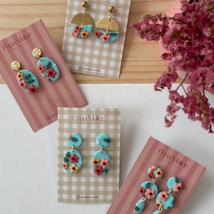 Puede incluir: Pendientes hechos a mano en varios diseños. Los pendientes presentan una base azul claro con detalles florales en rojo, naranja y rosa. Algunos pendientes tienen la parte superior en forma de semicírculo dorado. Los pendientes se muestran en tarjetas con la marca "Pepa Mulier".