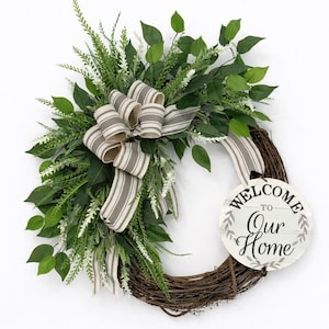 May include: A decorative grapevine wreath adorned with lush green foliage, white accents, and a striped bow. A round wooden sign with the text "WELCOME to Our Home" is attached, creating a welcoming home decor piece.