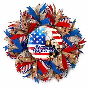 May include: A patriotic wreath featuring red, white, and blue ribbons and bows. The wreath has a central circular design with the American flag, a bald eagle, and the text "God Bless America."
