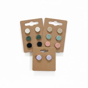 May include: Several pairs of stud earrings in a variety of colours, each featuring a striped pattern. The earrings are presented on small, brown cardboard earring cards. Colours include white, green, black, beige, blue, pink, and purple.