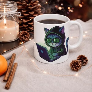 Pode incluir: Caneca de cerâmica branca com um gato de desenho animado usando um capuz e óculos de sol, usando um laptop. O gato é azul e verde. A caneca contém um líquido escuro. O fundo inclui uma vela acesa, pinhas e paus de canela.