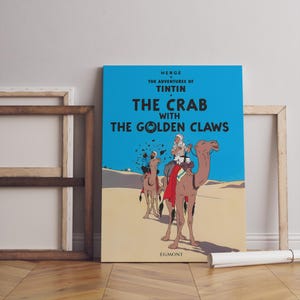 May include: A framed poster of "The Adventures of Tintin: The Crab with the Golden Claws" by Herg&eacute;. The poster features a blue background with the title in white and yellow. The illustration shows characters riding camels in a desert setting.