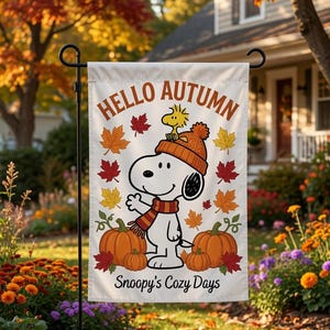Könnte beinhalten: Eine rechteckige Gartenflagge mit weißem Hintergrund, dem Text HELLO AUTUMN oben und Snoopy's Cozy Days unten. Das Motiv zeigt einen Cartoon-Hund mit einer orangefarbenen Strickmütze und einem Schal, umgeben von Kürbissen und herbstlichen Blättern.