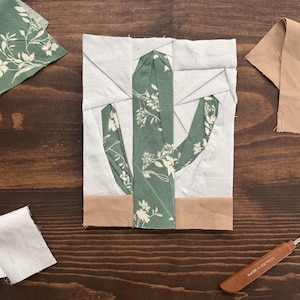 May include: A rectangular fabric quilt block featuring a pieced cactus design. The cactus is made from sage green fabric with a white floral pattern, set against a white background with a solid tan fabric strip at the bottom base of the block.