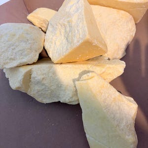 May include: A collection of irregularly shaped, pale yellow cocoa butter chunks. The pieces vary in size and have a rough, textured surface. The cocoa butter is displayed on a brown surface, suggesting a natural or artisanal product.