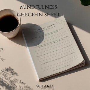 Könnte beinhalten: Ein cremefarbenes Journal mit dem Text "MINDFULNESS CHECK-IN SHEET" und dem Markennamen "SOLARIA" liegt neben einer Tasse Kaffee. Die Seiten des Journals sind mit Aufforderungen versehen. Die Kaffeetasse ist weiß und mit dunkler Flüssigkeit gefüllt.
