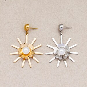 May include: A pair of stud earrings featuring a sunburst design with radiating metal spikes surrounding a central four-petaled flower shape. One earring is gold-toned and the other is silver-toned, each with a white pearl-like bead at the center.