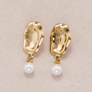 May include: A pair of gold-toned earrings featuring an irregular, molten-textured metal plate at the top. Each earring is finished with a single, round, white pearl drop suspended from a small circular jump ring attached to the base of the plate.