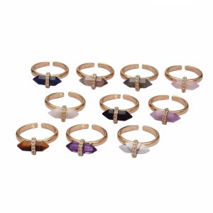 May include: A collection of ten adjustable gold-tone rings, each featuring a horizontal double-pointed gemstone in various colors including blue, brown, black, white, and purple, accented by a small vertical band of clear crystals.