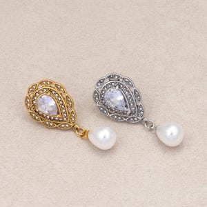 May include: Two teardrop-shaped earrings, one in gold tone and one in silver tone. Each earring features a central pear-cut clear stone surrounded by small pavé crystals, with a dangling white pearl drop attached to the bottom of the frame.