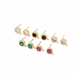 May include: A collection of ten gold-toned stud earring components, each featuring a circular bezel setting with a small loop at the base. The studs contain faceted stones in various colors including clear, black, pink, green, and magenta.