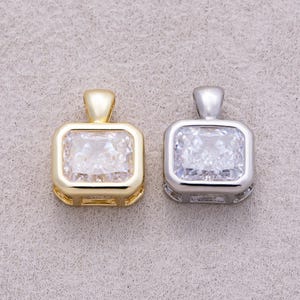 May include: Two rectangular pendants featuring large, clear, faceted stones set in metal frames. One pendant is finished in a warm gold tone, while the other is finished in a cool silver tone. Both pendants include a matching metal bail at the top.