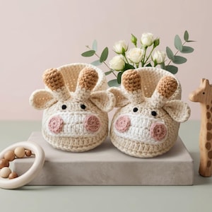 Op de afbeelding: Een paar crèmekleurige gehaakte babyslofjes in de vorm van giraffen. De slofjes hebben bruine oren, roze wangen en zwarte ogen. Een houten giraffenspeelgoed en een houten bijtring staan ook op de foto.