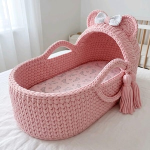 Può includere: Una culla per neonati all'uncinetto rosa fatta a mano con cappuccio a forma di orecchie e fiocco bianco. La culla ha manici, nappe e un materasso fantasia. Il design è morbido e invitante. Misura circa 70 cm di lunghezza.