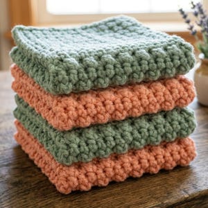 Peut inclure: Une pile de quatre débarbouillettes faites au crochet. Deux sont vert sauge et deux sont de couleur pêche douce. Les débarbouillettes ont une bordure texturée en relief et une forme carrée. La pile est sur une surface en bois, avec une fenêtre et des fleurs en arrière-plan.