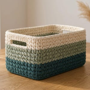 May include: A rectangular crochet basket in cream, green, and teal. The basket has a handle on each side and is made of thick yarn. The color scheme is arranged in horizontal stripes. This storage basket is suitable for home organization.
