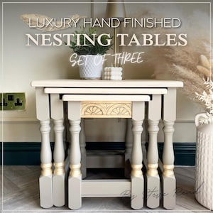 May include: A set of three nesting tables with light grey rectangular tops and turned legs featuring gold-painted accents at the base. The front apron of the smallest table displays a carved semi-circular fan pattern. Text reads: LUXURY HAND FINISHED NESTING TABLES SET OF THREE.