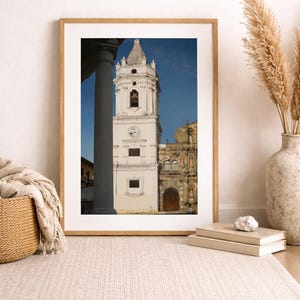 May include: Framed photograph of a white church tower with a clock and bell tower, set against a blue sky. The image is in a light wood frame, with a neutral rug and decorative vase in the foreground.