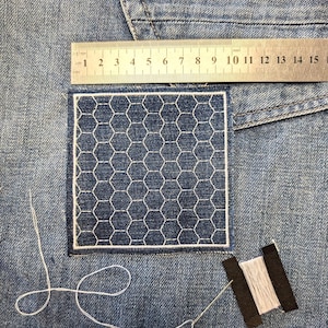 May include: Close-up of denim fabric with a white embroidered patch in a honeycomb design. A metal ruler shows measurements in centimetres. A needle and thread are next to the patch, along with a spool of white thread.