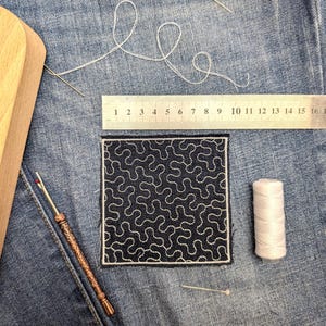 May include: A square navy blue fabric patch with a white swirling design is shown on denim. A ruler, pins, white thread, a seam ripper, and a spool of white thread are also visible. The ruler measures up to 16 cm.