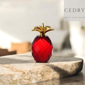 Op de afbeelding: Een facetgeslepen, robijnrood glazen decoratief object in de vorm van een ananas met een goudkleurige bovenkant. Het object wordt weergegeven op een marmeren oppervlak. De tekst "CEDRY LUXURY GLASS DECOR" staat op de achtergrond.
