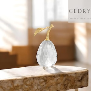 Op de afbeelding: Een heldere, facetgeslepen glazen perensculptuur met een goudkleurige steel en blad. Het peervormige ornament wordt getoond op een marmerachtig oppervlak. De tekst "CEDRY LUXURY GLASS DECOR" staat rechtsboven.