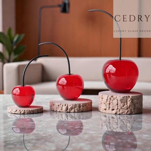 May include: Three decorative glass cherry sculptures in varying sizes. Each cherry is a vibrant red with a glossy finish, featuring a slender black stem. The cherries rest on cork bases, reflecting on the surface below. The text "CEDRY LUXURY GLASS DECOR" is visible.