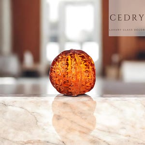 Puede incluir: Un objeto decorativo de vidrio naranja, con forma de fruta segmentada, sobre una superficie de mármol. El vidrio tiene una superficie texturizada y refleja la luz. El texto "CEDRY LUXURY GLASS DECOR" es visible en el fondo.