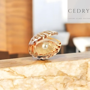 May include: A decorative glass seashell with a pearl inside. The shell is a translucent amber color, and the pearl is white. The image also includes the text "CEDRY LUXURY GLASS DECOR".