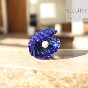 May include: A blue glass seashell with a white pearl inside. The shell is open, revealing the pearl. The glass is transparent, allowing light to pass through. The text "CEDRY LUXURY GLASS DECOR" is in the background.