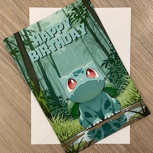 May include: A rectangular greeting card featuring a light blue Bulbasaur character with red eyes and a green bulb on its back, sitting in a forest with the text HAPPY BIRTHDAY in light blue letters. The card is placed on a plain white envelope.