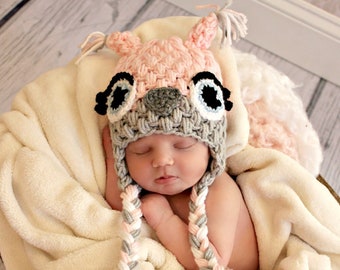 May include: A newborn baby wearing a pink and gray crocheted owl hat with braided ties. The baby is sleeping in a white blanket inside a wooden crate.
