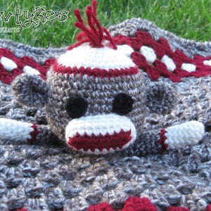 May include: A crocheted baby blanket with a gray and white pattern and red accents. A crocheted monkey toy with red, white, and gray yarn is on top of the blanket. The monkey has a white face with a red mouth and black eyes.