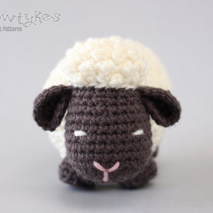 May include: A crocheted stuffed sheep toy with a white fleece and a brown face. The sheep has black eyes and a pink nose.