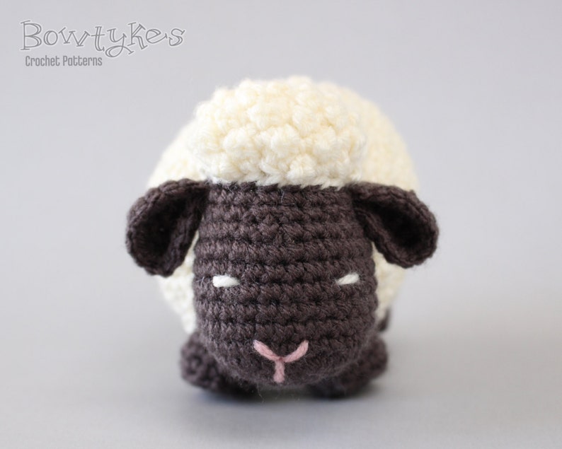 May include: A crocheted stuffed sheep toy with a white fleece and a brown face. The sheep has black eyes and a pink nose.