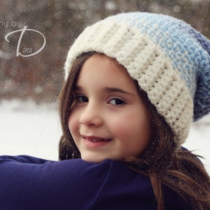May include: A young girl wearing a white and blue crocheted hat with a white and blue striped pattern. She is smiling and looking at the camera. The background is a snowy forest.
