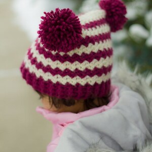 Puede incluir: Un gorro de punto de rayas rosa y blanco con dos pompones burdeos en la parte superior. El gorro lo lleva puesto un niño que lleva una chaqueta rosa con ribete de piel blanca.