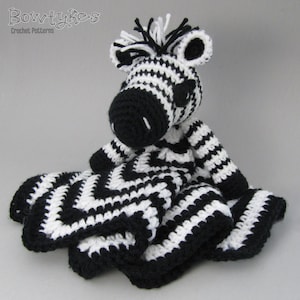 Pode incluir: Pelúcia de zebra em crochê preto e branco com um cobertor combinando. A zebra tem um nariz preto e olhos pretos. O cobertor é um padrão chevron.